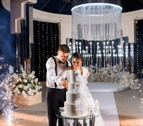 Front view happy married couple loving man and woman cutting big white cake on wedding ceremony at night. Standing near well-decorated luxury altar with flowers and sparklers. Special event